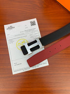 Black leather belt with gold-tone H buckle and red underside, shown beside paperwork.