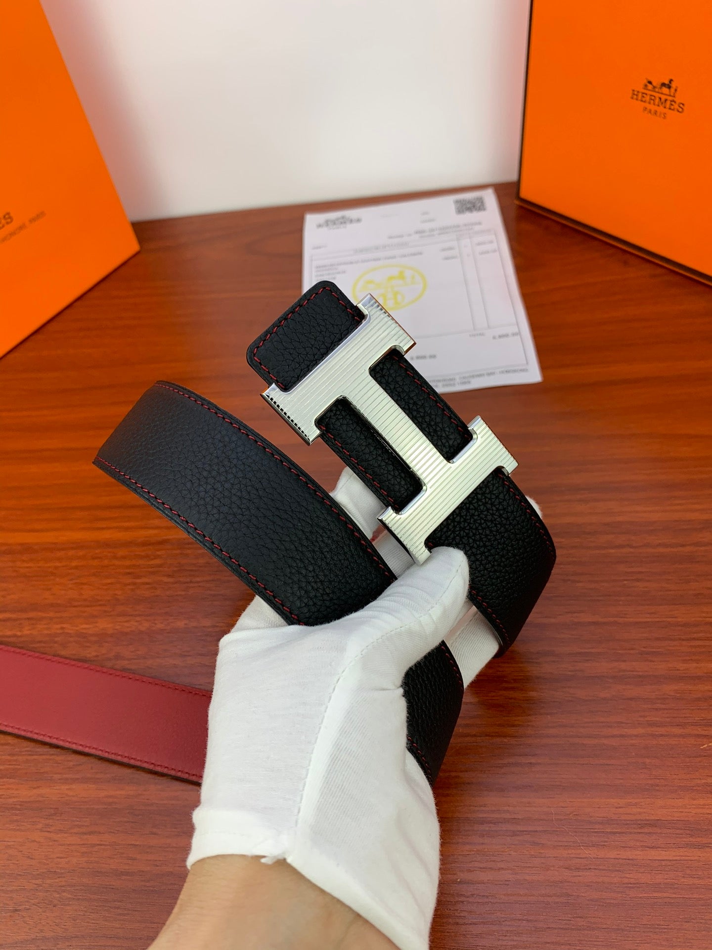 Black leather belt with gold-tone H buckle and textured finish, held by white glove.
