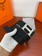 Black leather belt with gold-tone H buckle and textured grain, held in white glove.