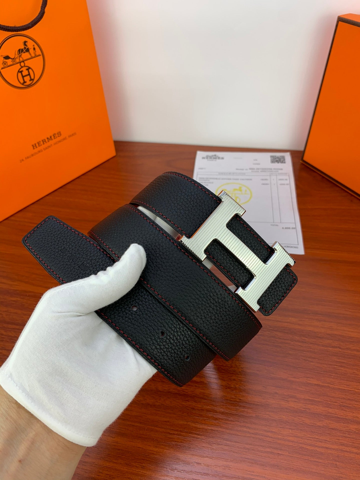 Black leather belt with gold-tone H buckle and textured grain, held in white glove.