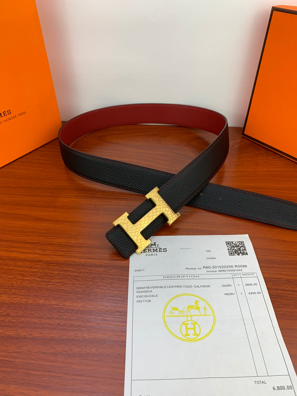 Black textured leather belt with gold-toned H buckle, laid on tabletop with card