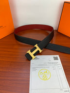 Black textured leather belt with gold-toned H buckle, laid on tabletop with card