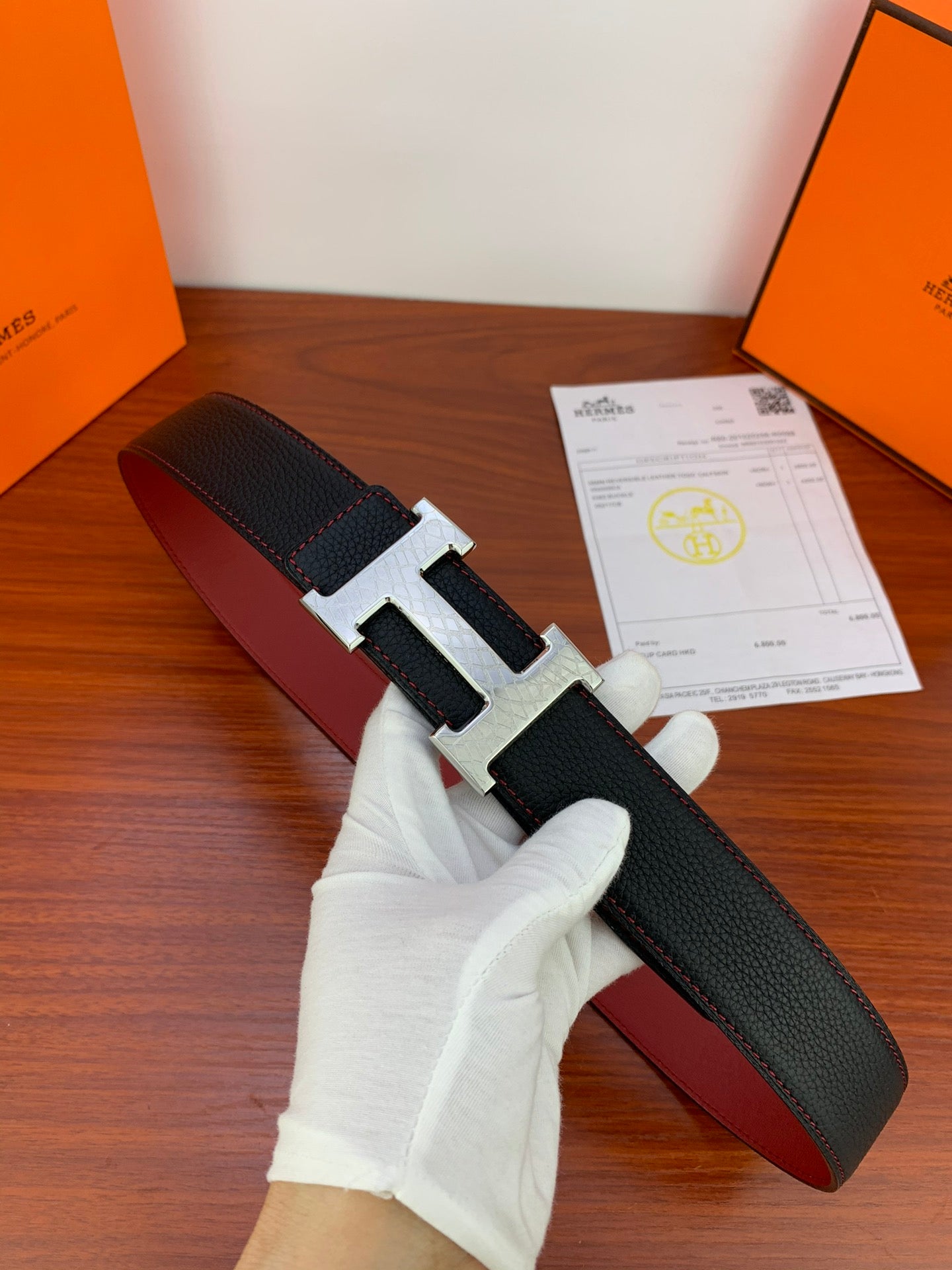 Black textured leather belt with silver-toned H buckle, held by white-gloved hand