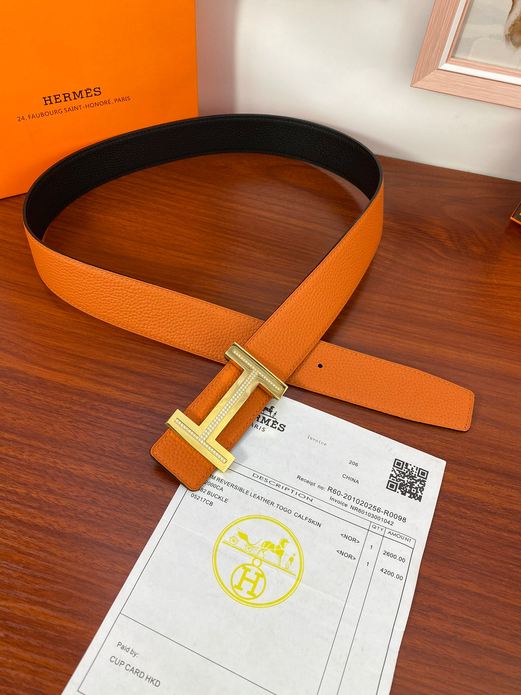 Reversible leather belt with orange strap and silver H buckle lying flat