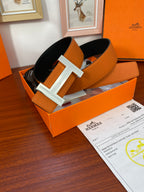 Reversible leather belt with orange strap and silver H buckle, shown laid in an open box