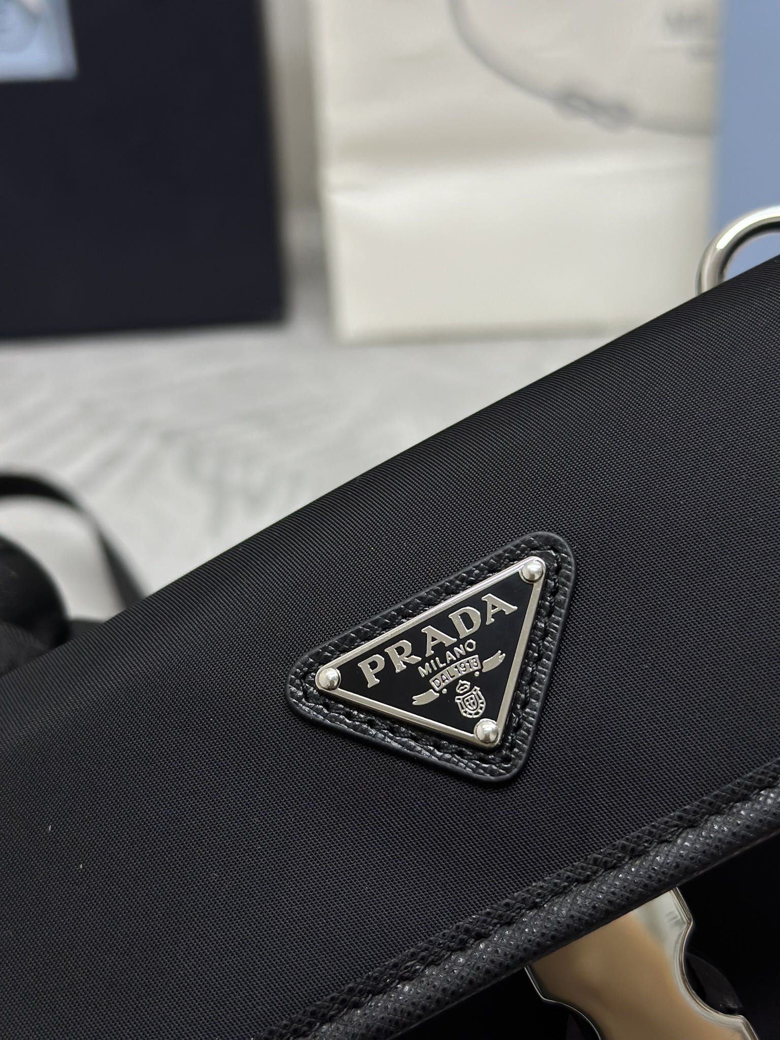 Prada bag front logo patch with silver-tone metal frame, dark textured surface