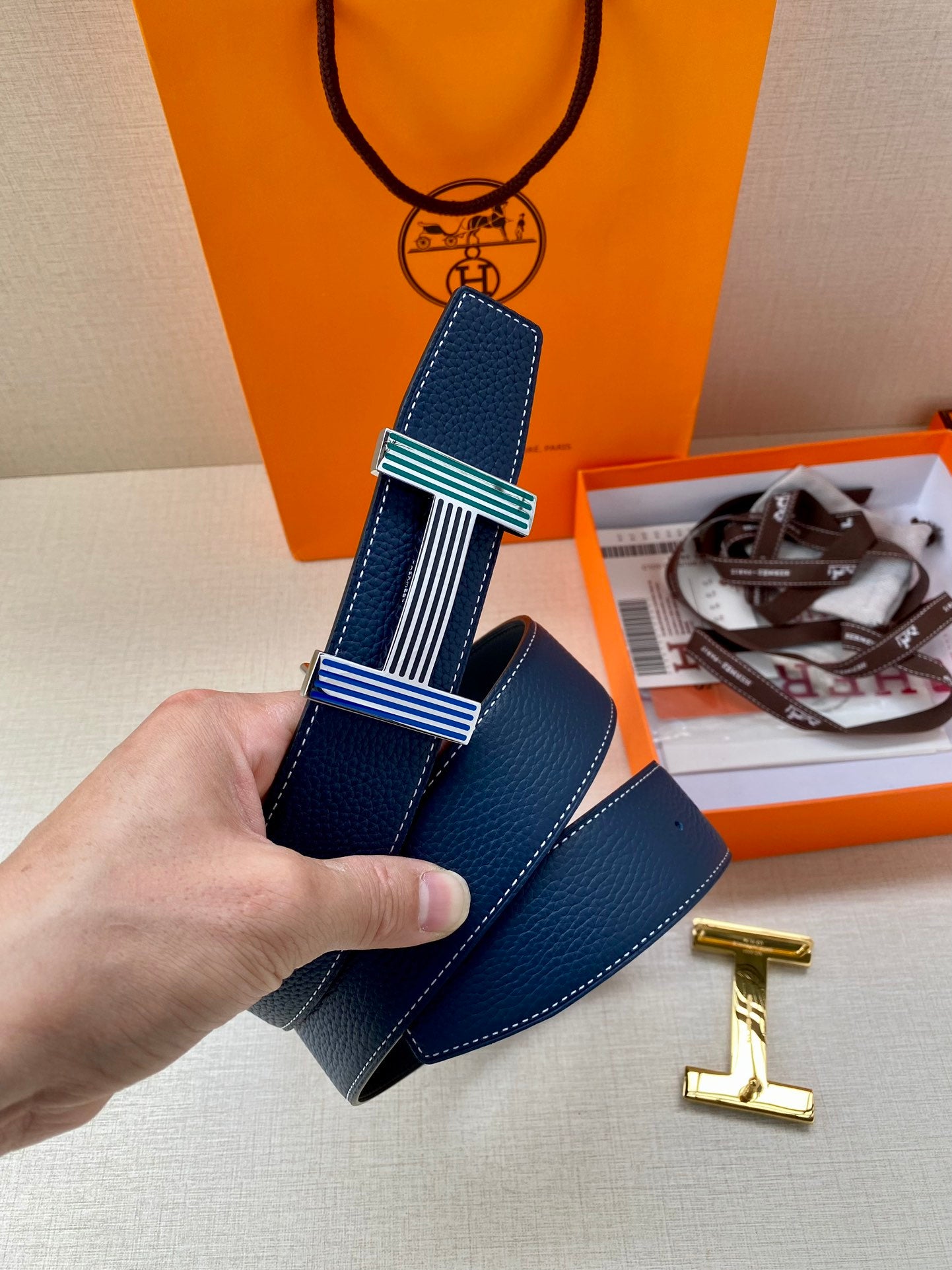 Blue leather belt with striped buckle and gold-toned H clasp, held up.