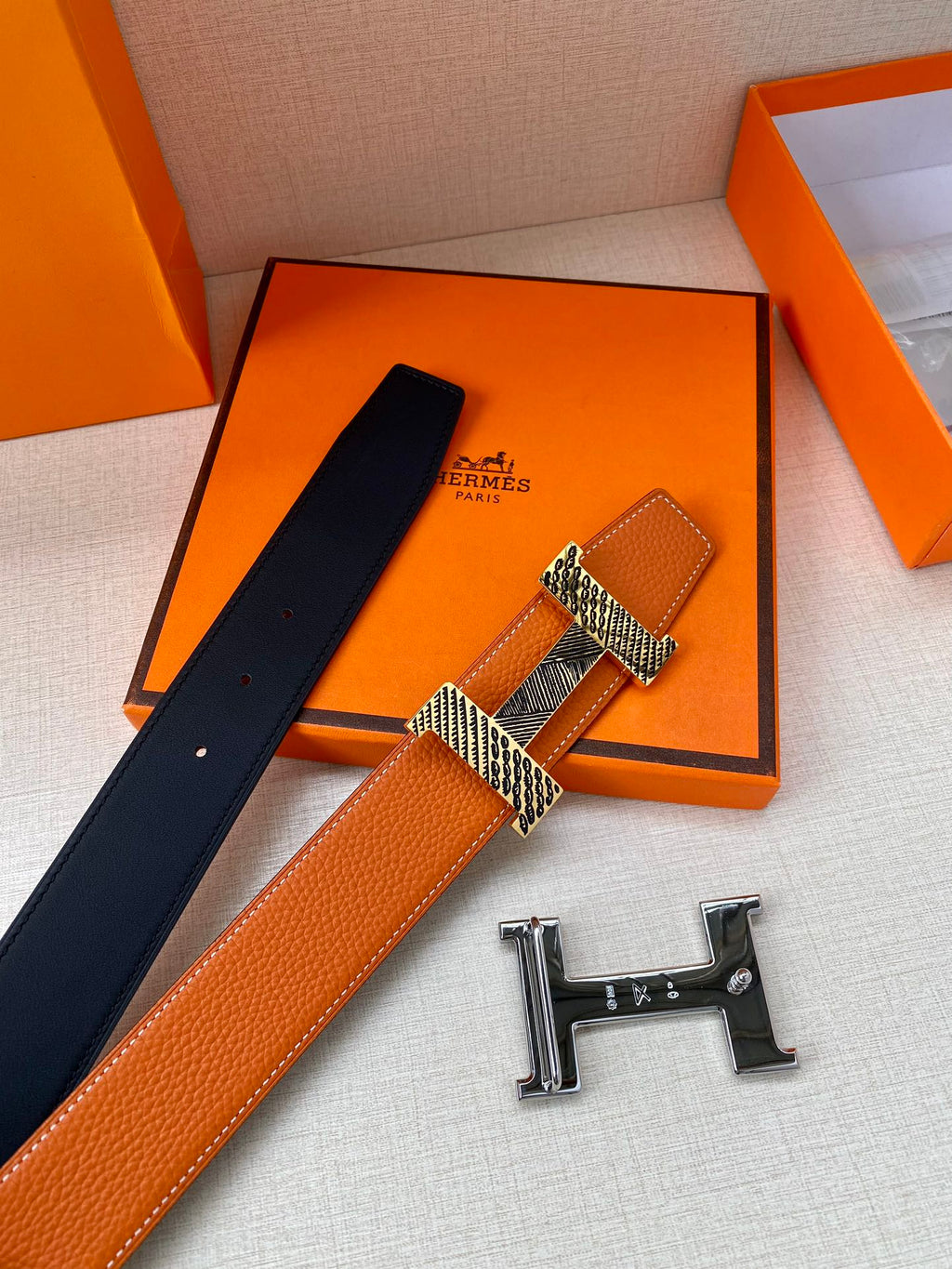 Orange leather belt with navy backing and gold-tone H buckle, shown with extra pieces
