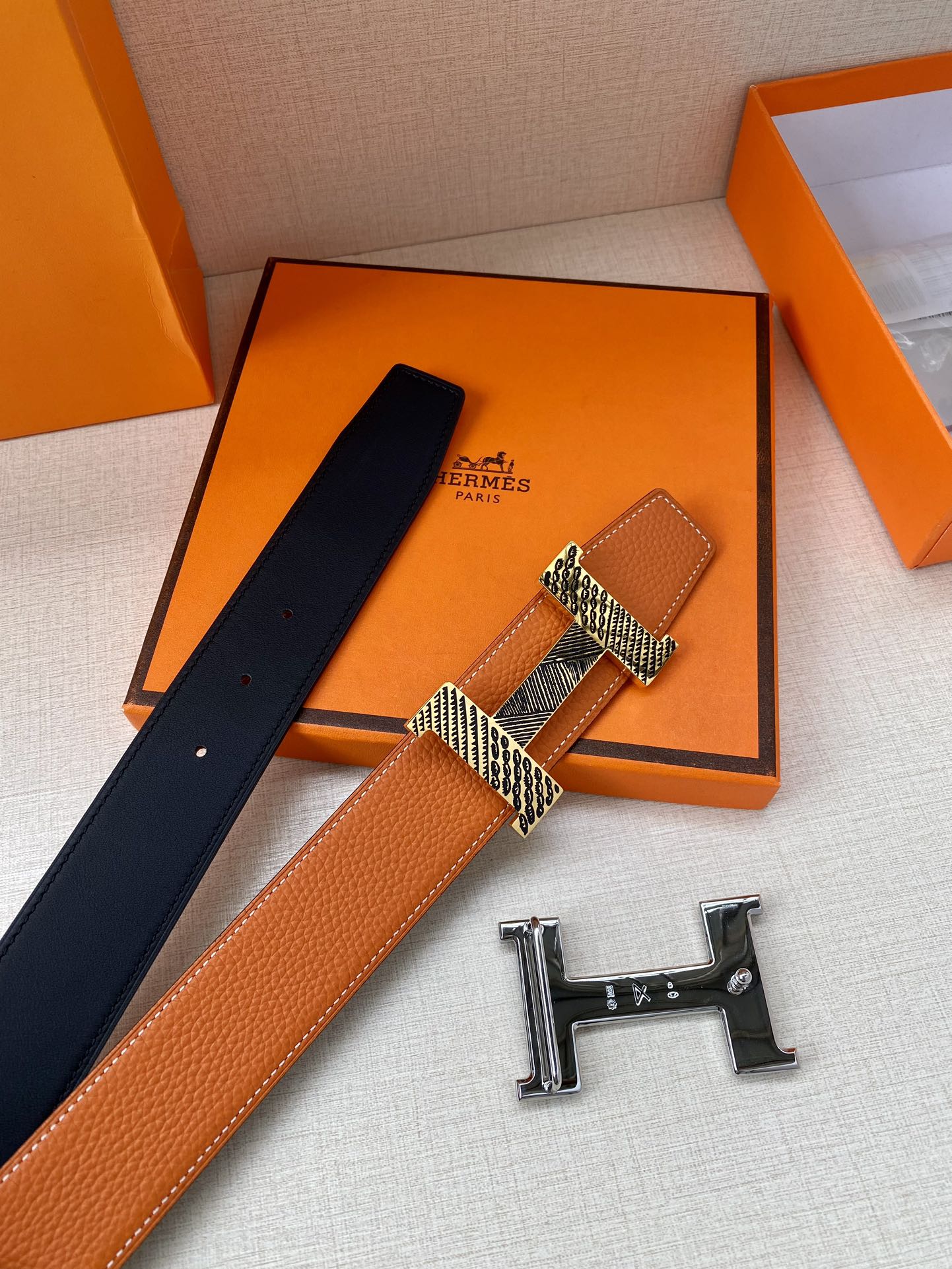 Orange leather belt with navy backing and gold-tone H buckle, shown with extra pieces