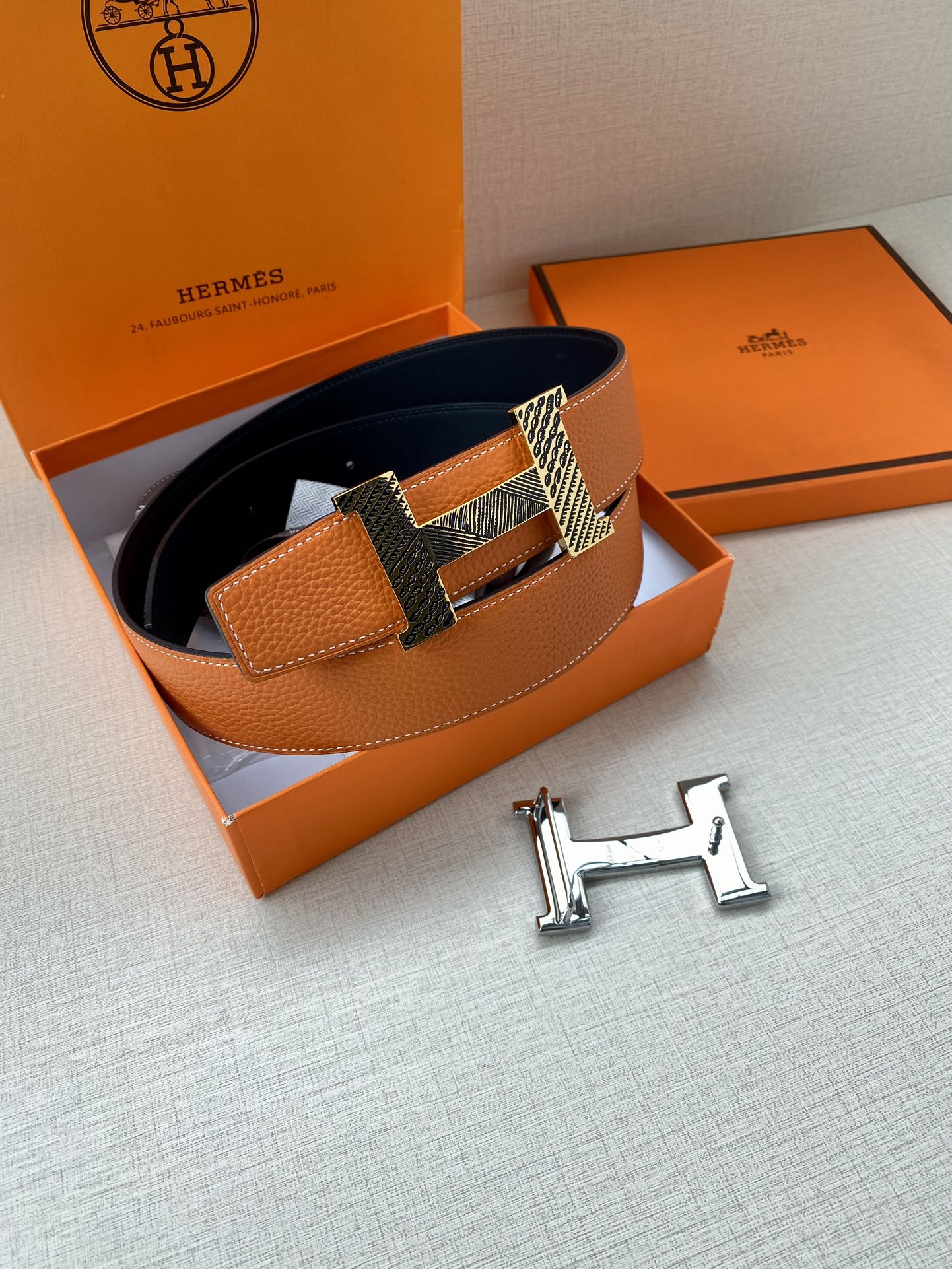 Orange leather belt with navy interior, textured buckle, and gold-tone H hardware in box