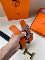 Orange leather belt with textured keeper and gold-tone H buckle, held in hand