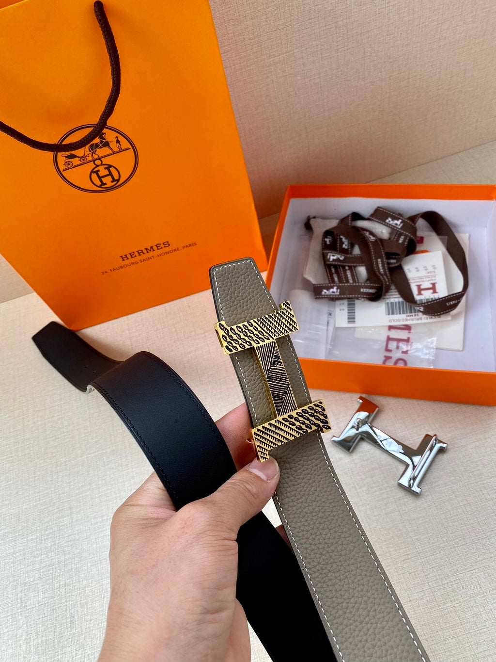 Hermes belt held in hand, gray strap with woven keeper and gold-toned H buckle