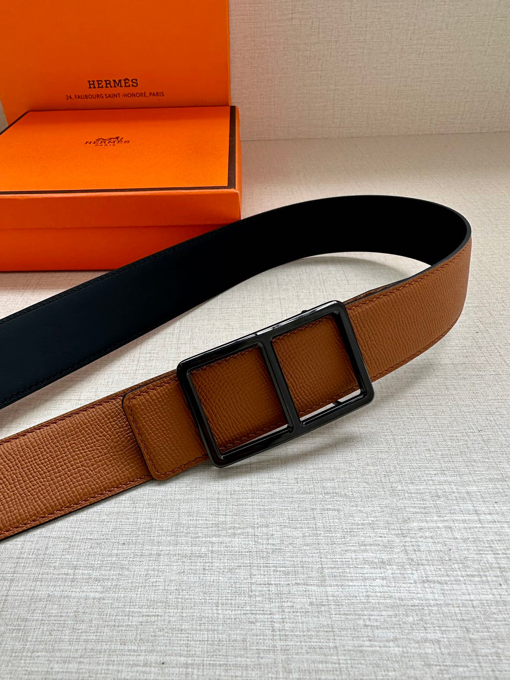 Hermes belt, tan strap with textured finish and dark square buckle, angled view