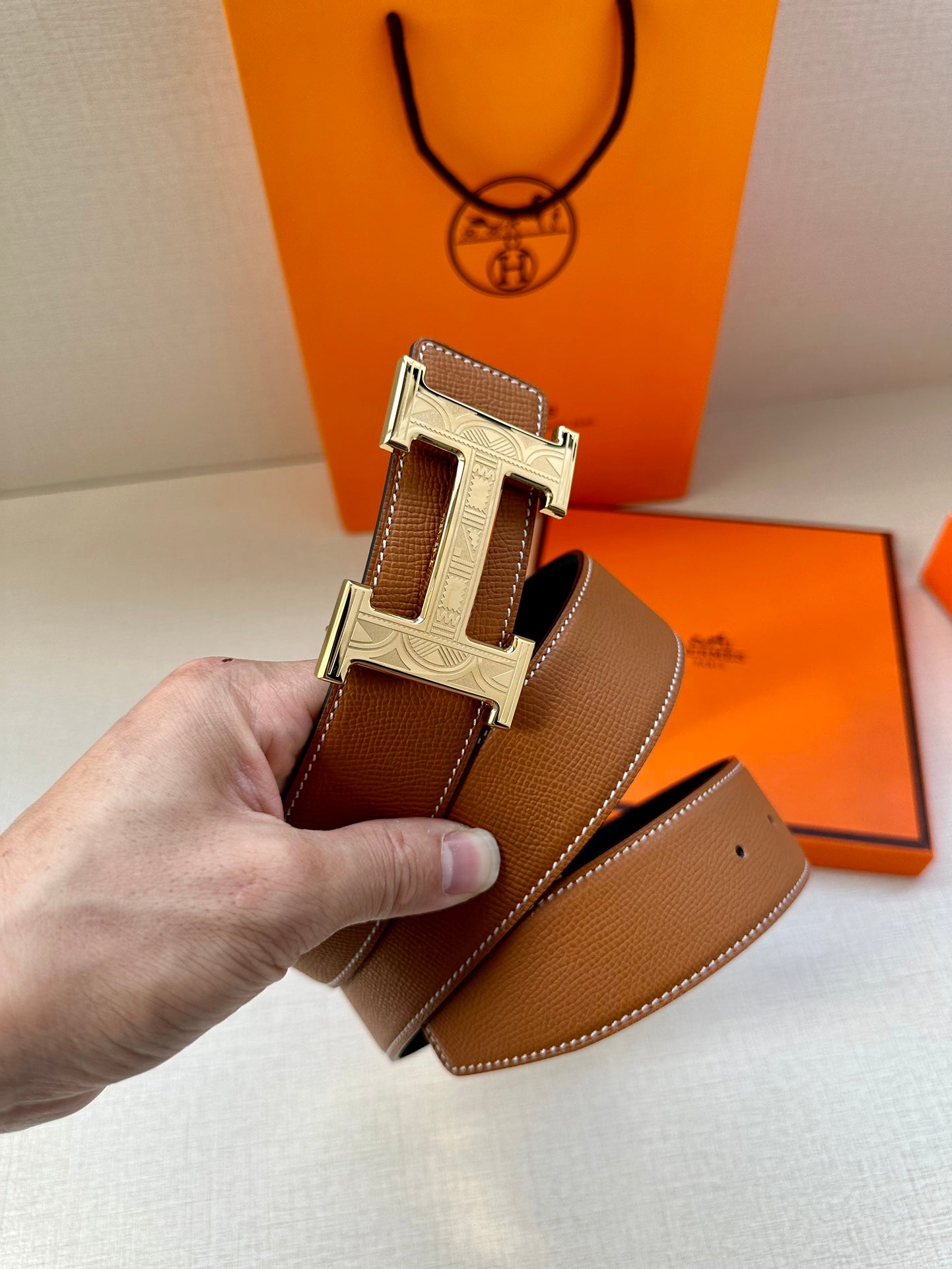 Tan leather belt with metal H buckle, held in hand with strap underside visible