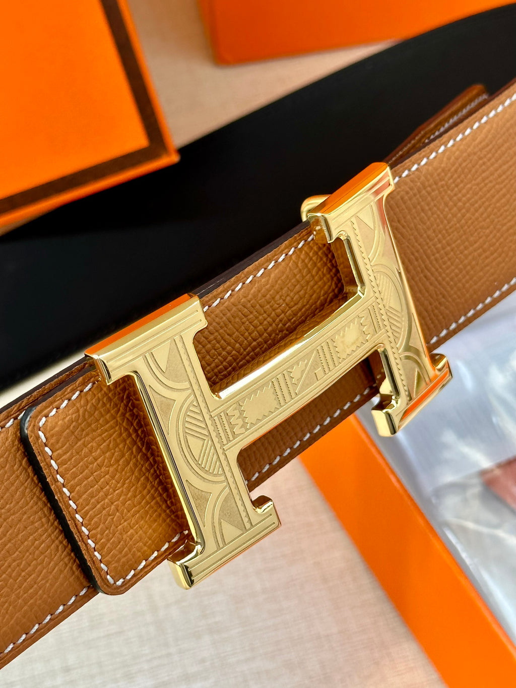 Tan leather belt with metal H buckle close-up, showing stitched strap end