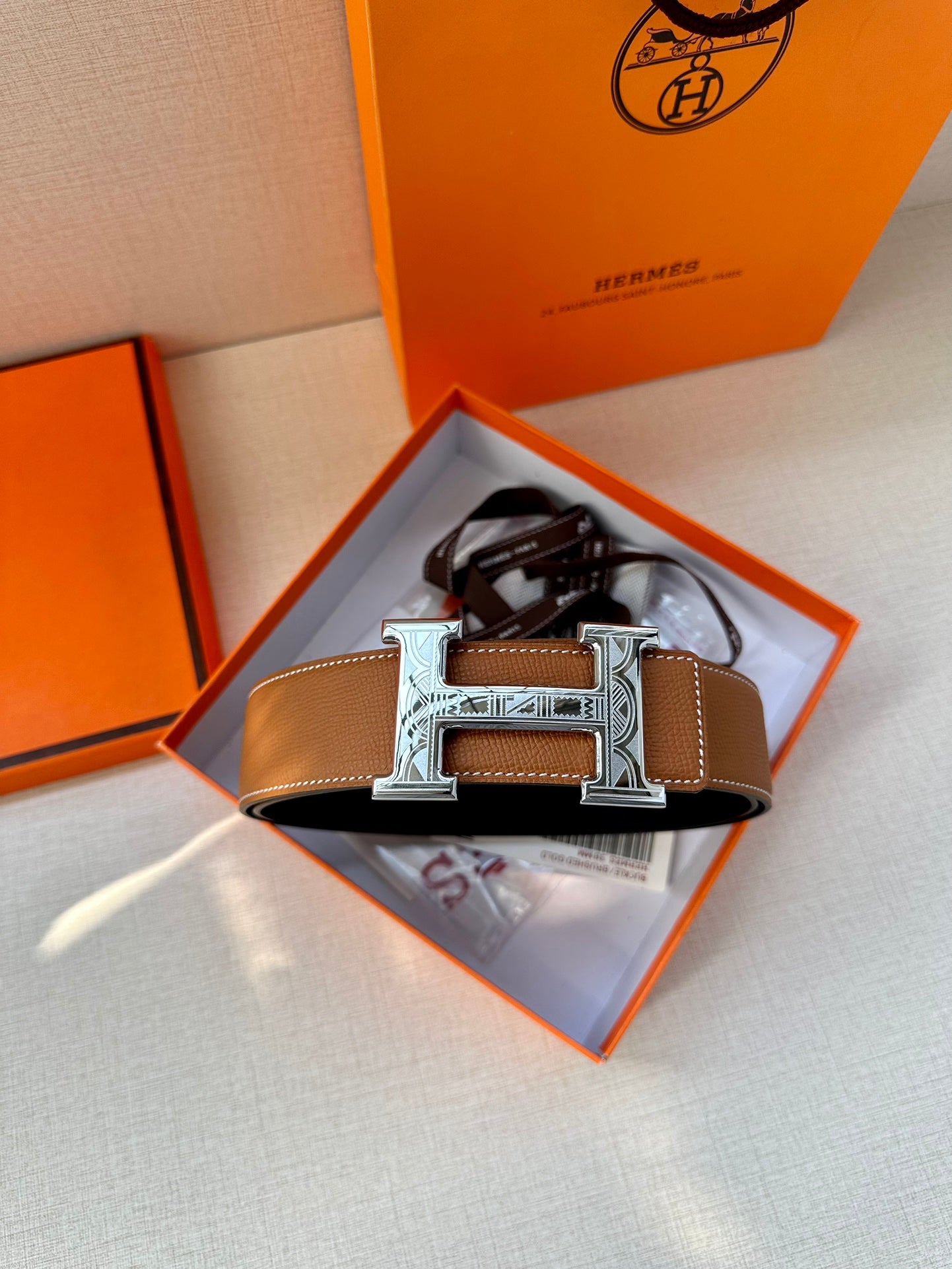 Tan leather belt with metal H buckle displayed in an open box