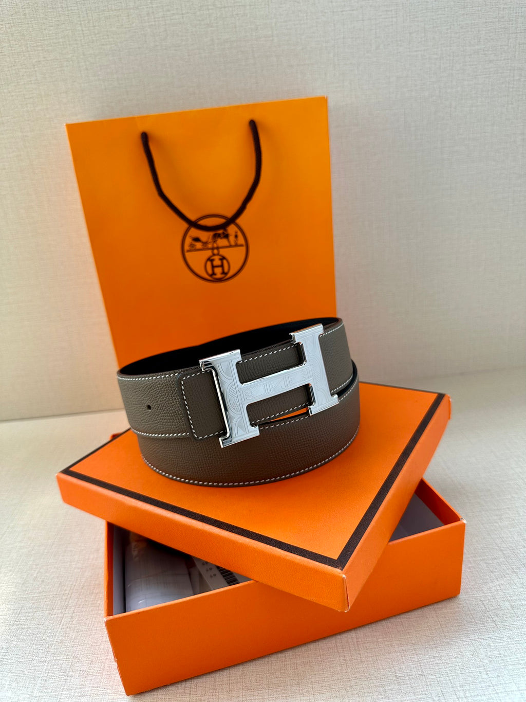 Leather belt with textured gray strap and silver-tone H buckle on a stand.