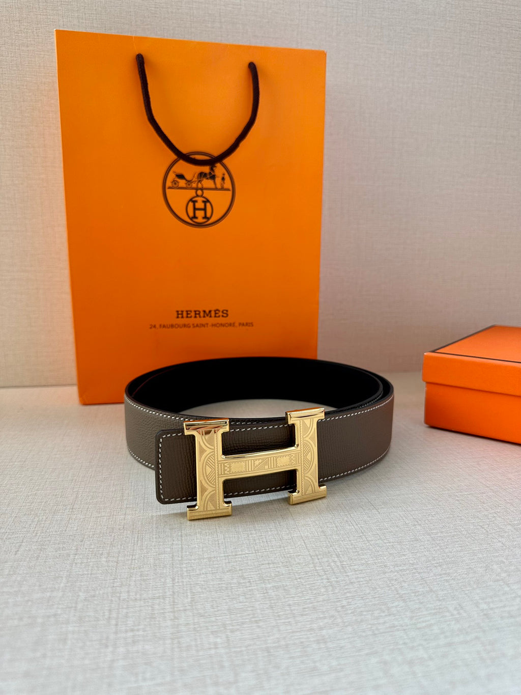 Leather belt with textured gray strap and gold-tone H buckle, laid flat.