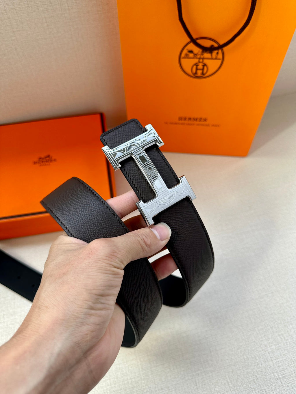 Black leather belt with silver-tone H buckle, side view held in hand.