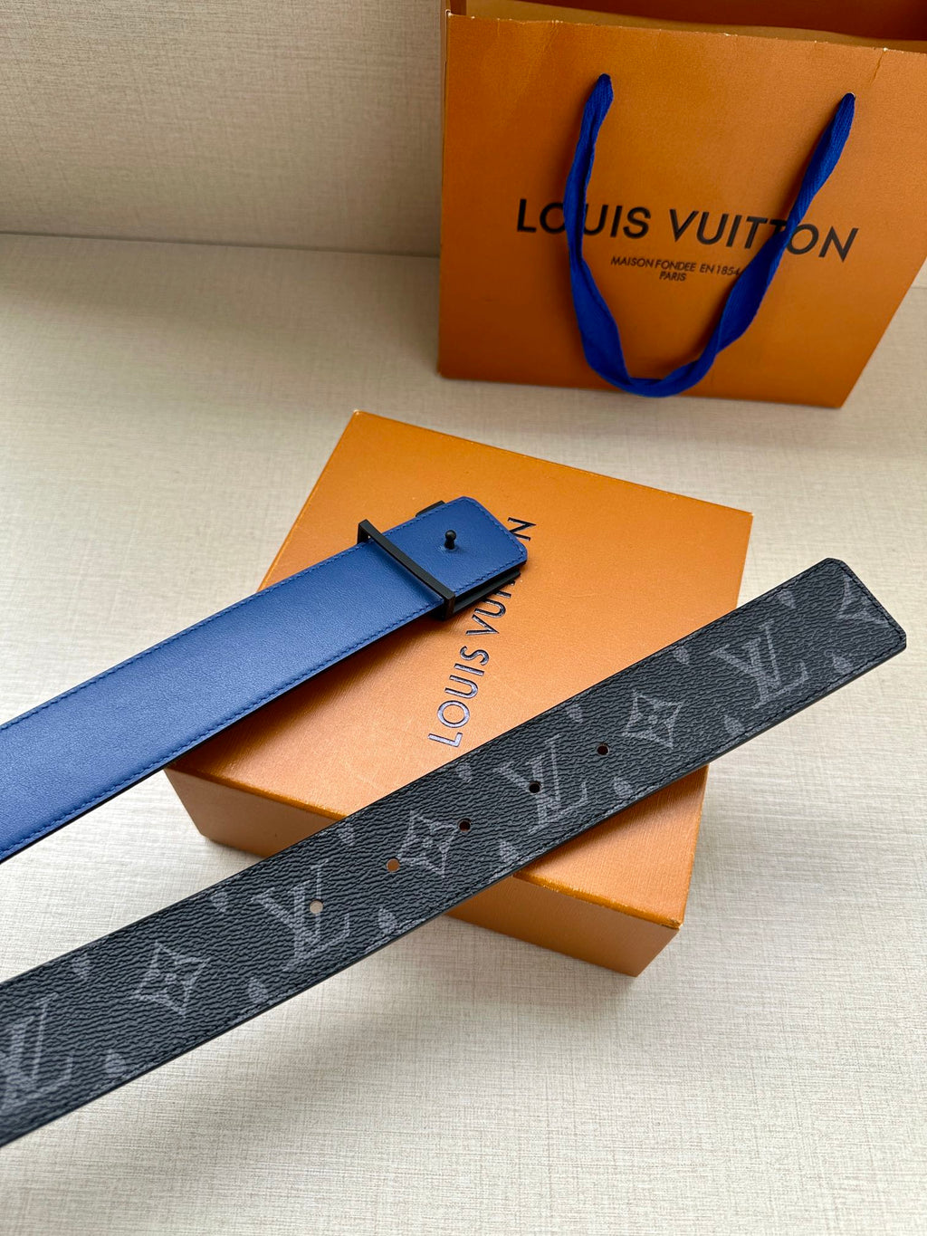 Louis Vuitton monogram belt with dark blue strap and gold-tone buckle on orange boxes.