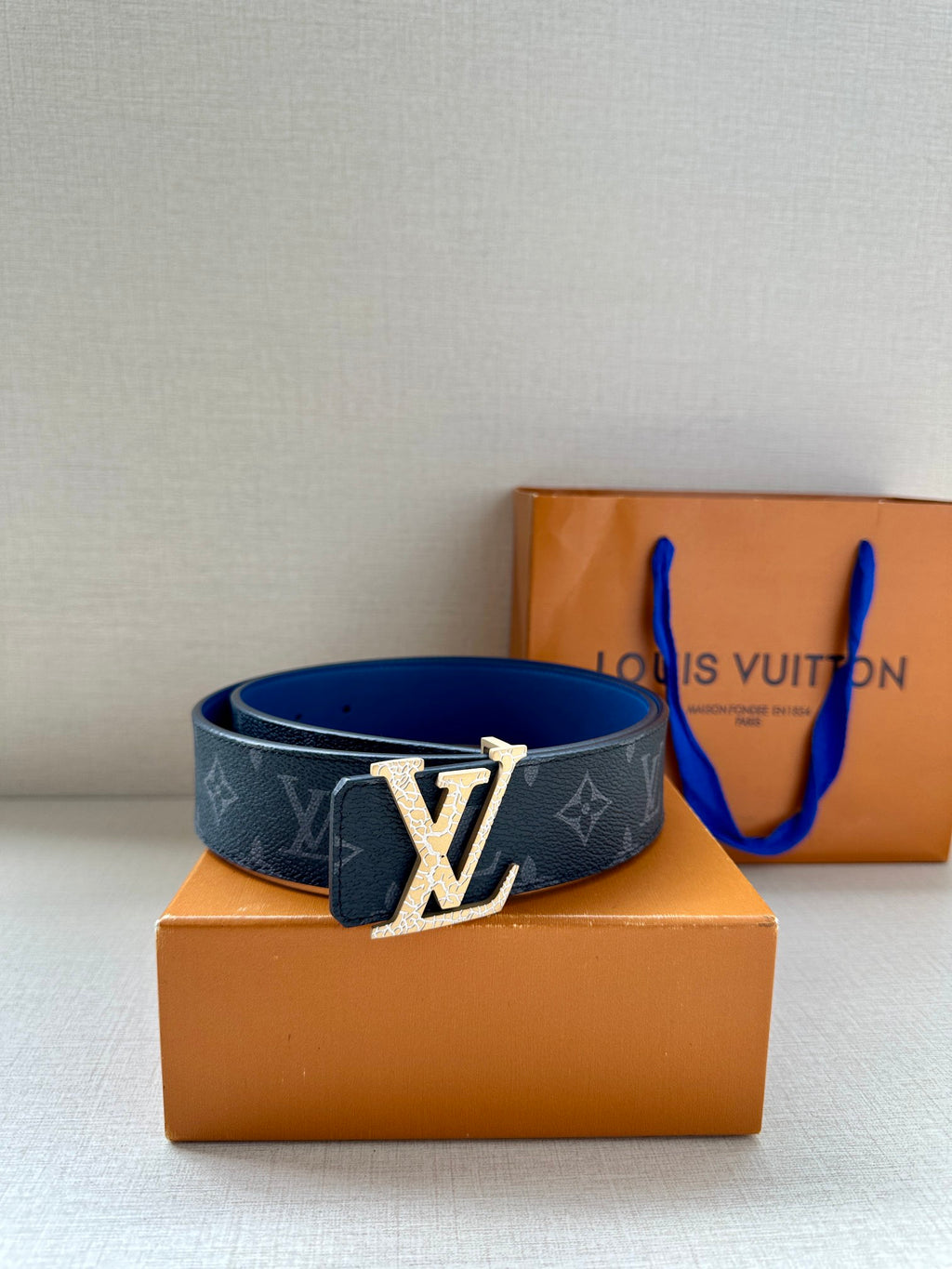 Louis Vuitton monogram belt with gold-tone LV buckle and dark blue strap on box.