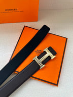 Hermes belt on orange box: dark strap with ornate silver H buckle and stitching.