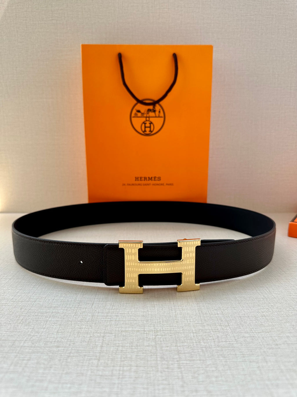 Hermes belt with dark strap and gold H buckle, laid flat with shopping bag behind.