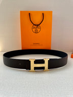 Hermes belt with dark strap and gold H buckle, laid flat with shopping bag behind.