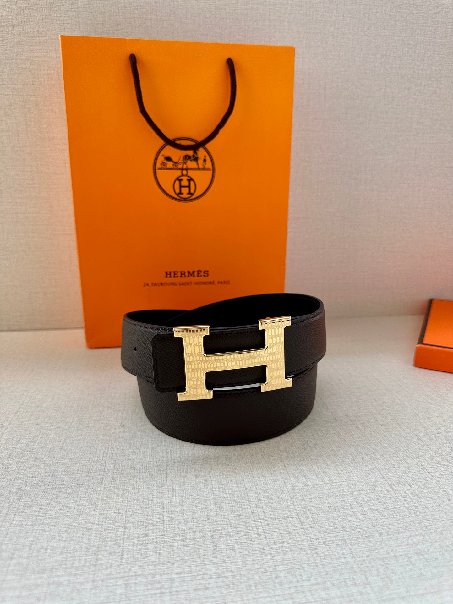 Hermes belt with dark strap and gold H buckle, laid flat beside gift boxes.