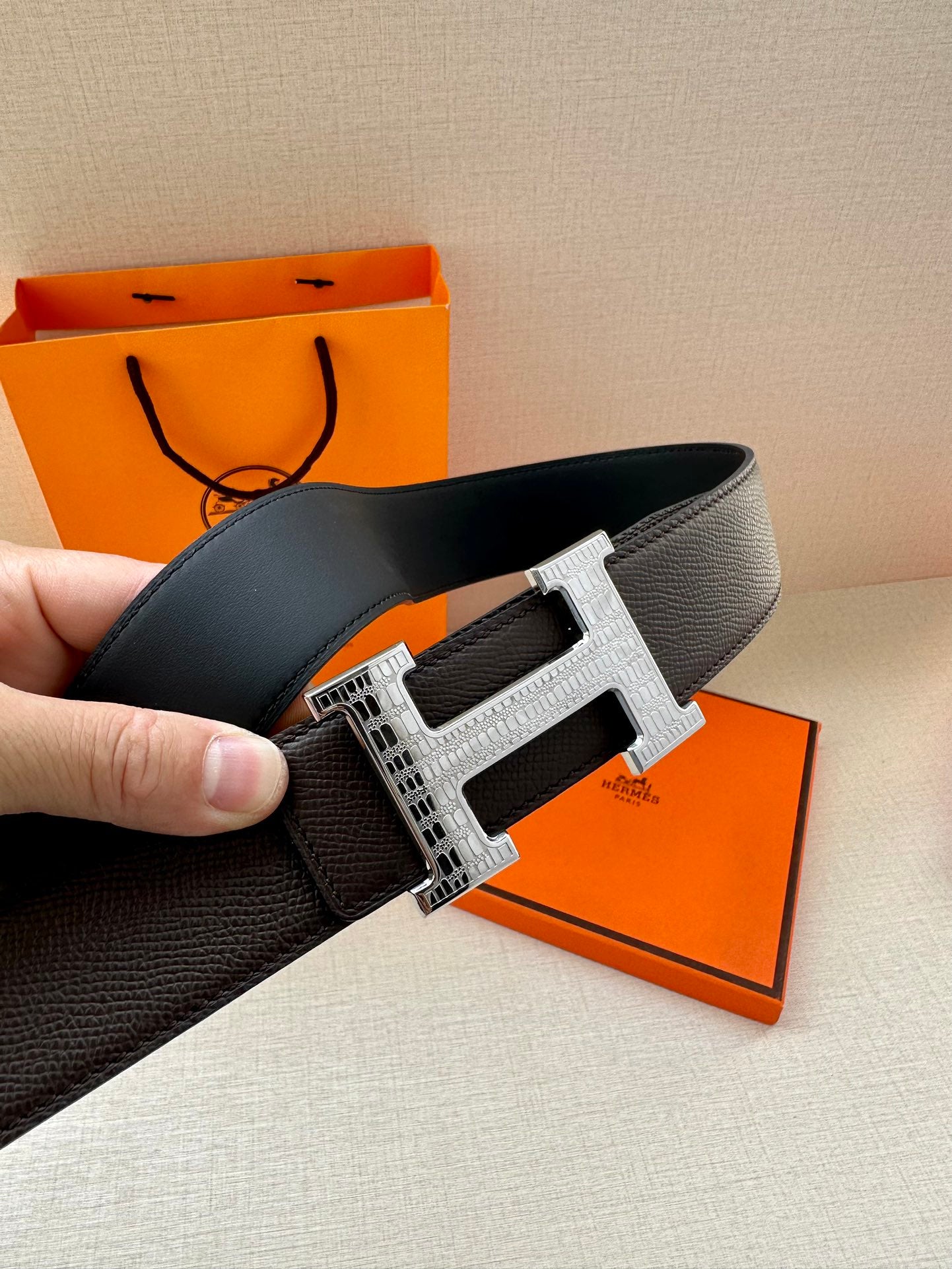 Hermes belt close-up with textured dark strap and silver H buckle on a hand.