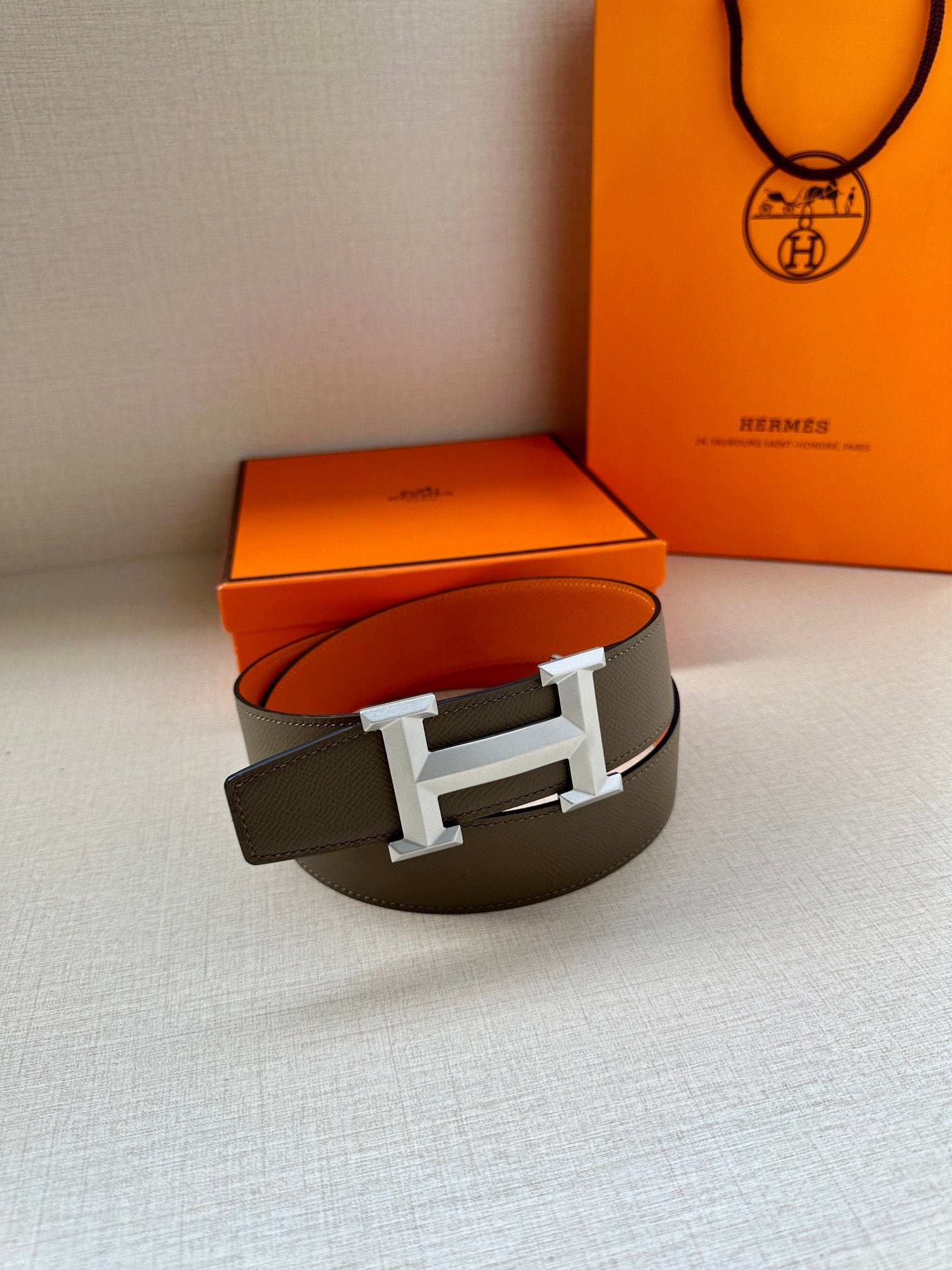 Leather belt with textured dark gray strap and silver-tone H buckle, on box.