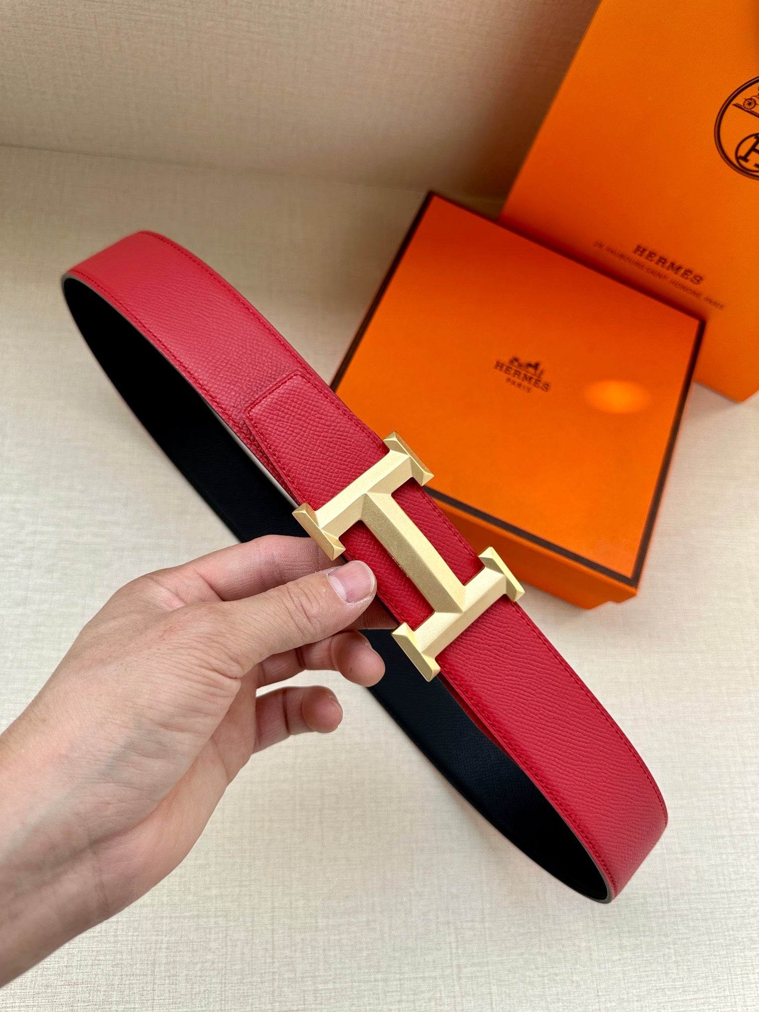 Red leather belt with gold H buckle, strap end held in a hand.