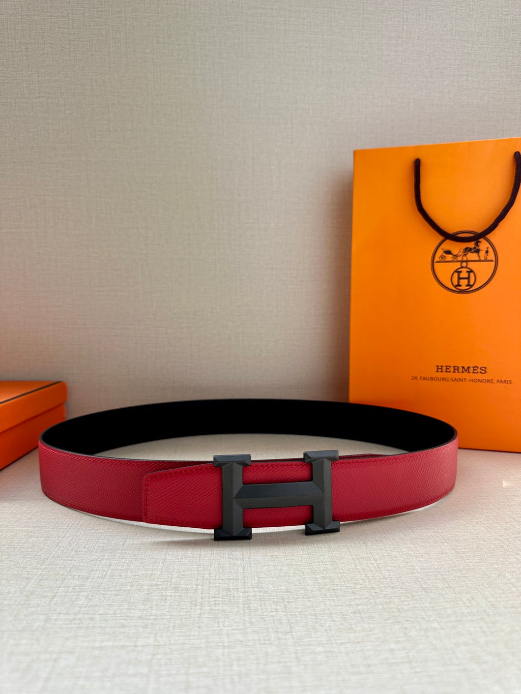 Red leather belt with dark H buckle, shown flat.