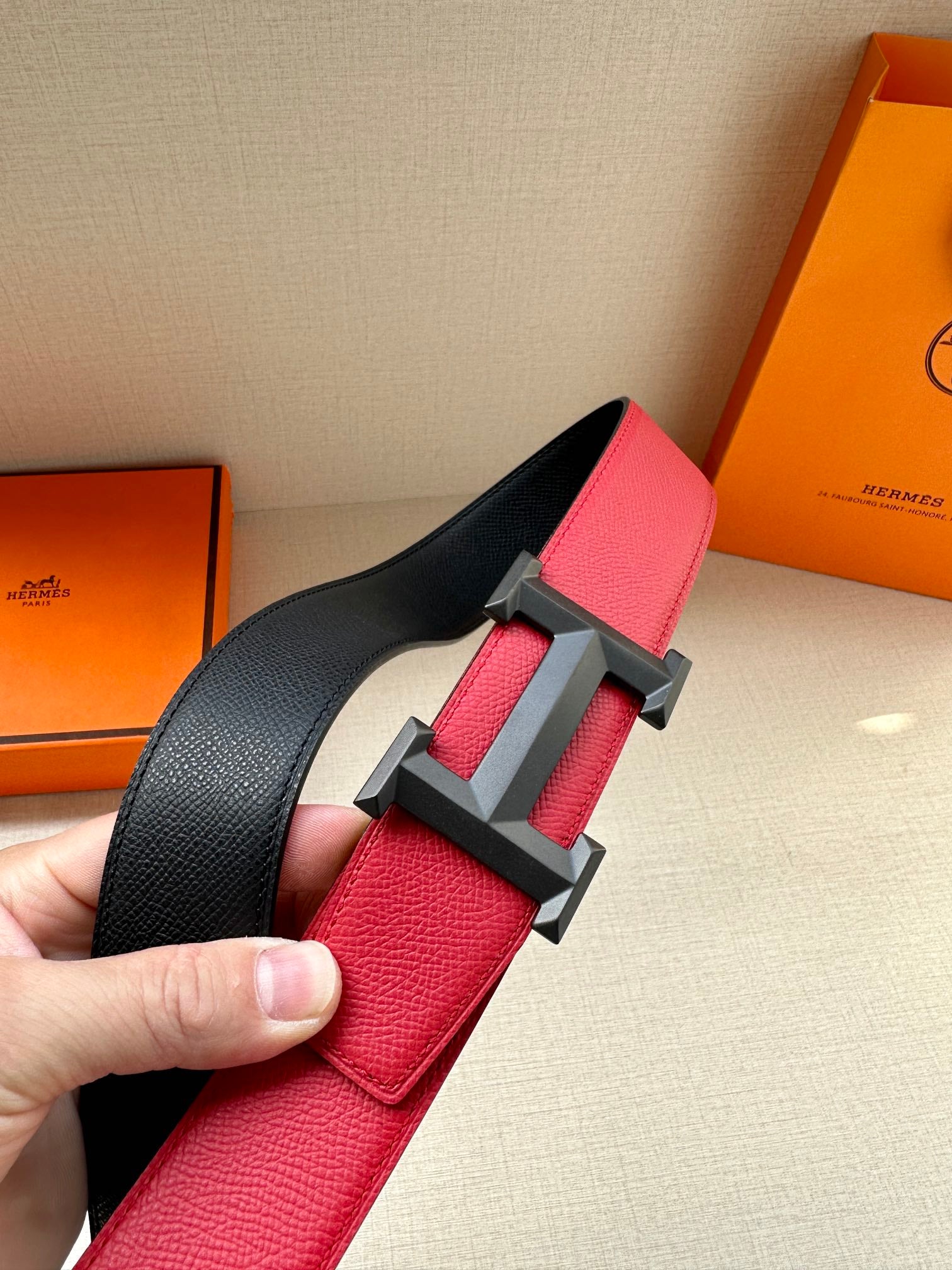 Red leather belt with dark H buckle and black strap section, close-up held in hand.