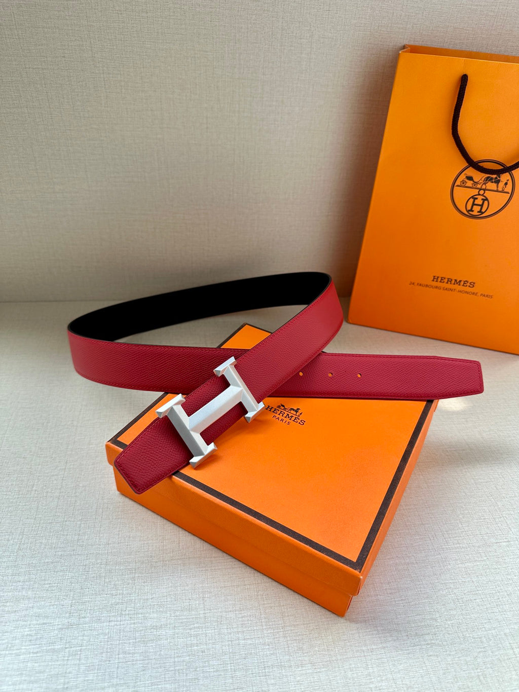 Red leather belt with black underside and metal H buckle, laid flat over a box.