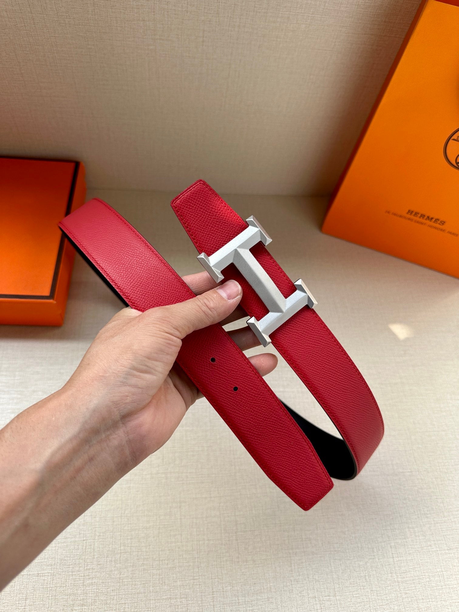Red leather belt with gold H buckle, hand holding the strap with hole detail.