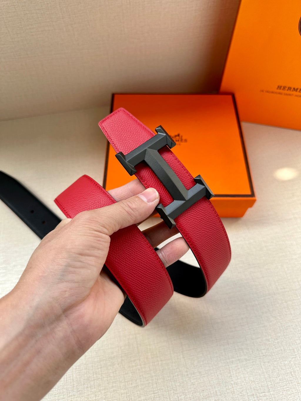 Red leather belt with dark H buckle, hand holding the strap end.