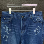 Blue denim pants close-up of waistband and front pockets with cross embroidery detail.