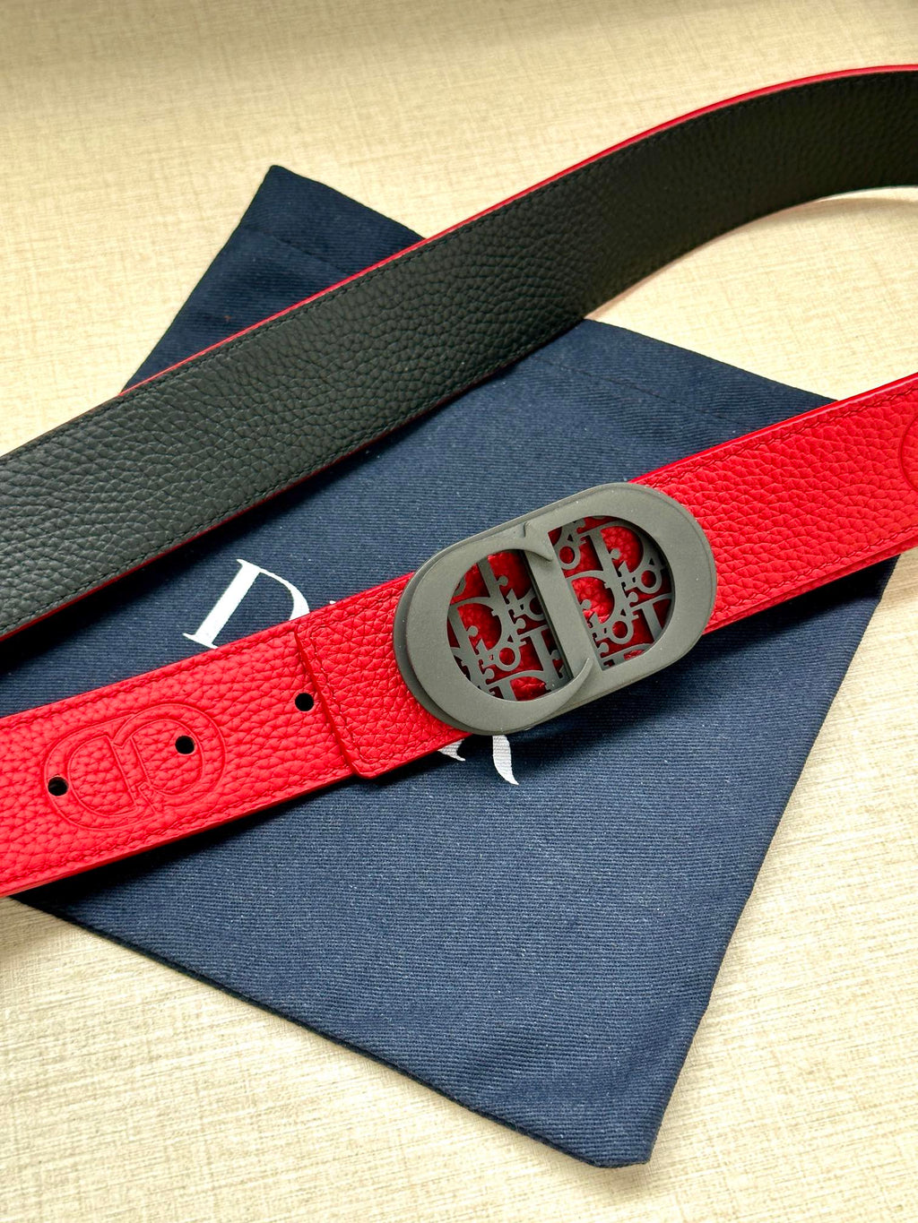 Red leather Dior belt with textured strap and gray oval patterned logo buckle.