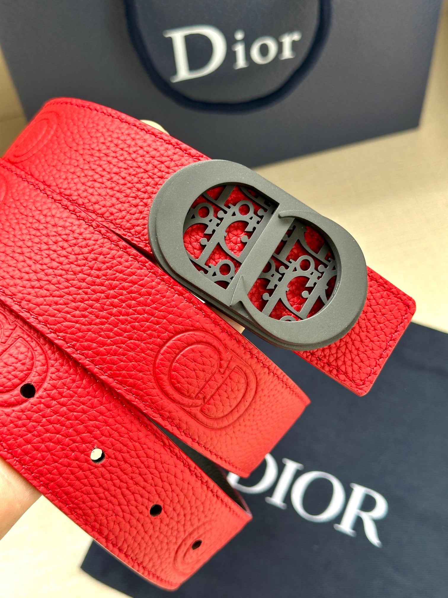 Red leather Dior belt close-up with gray oval buckle and embossed monogram on strap.
