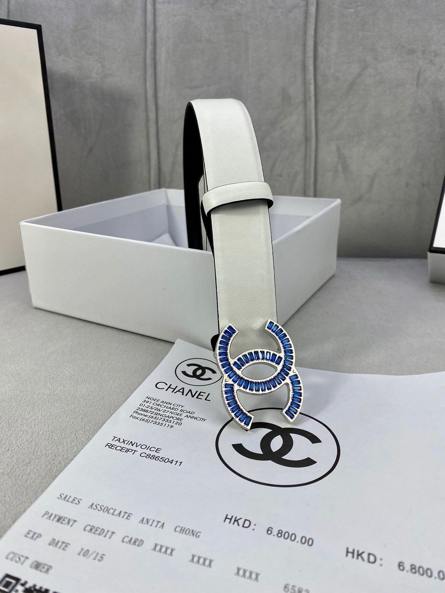 White leather belt with blue CC buckle and adjustable strap holes in box.