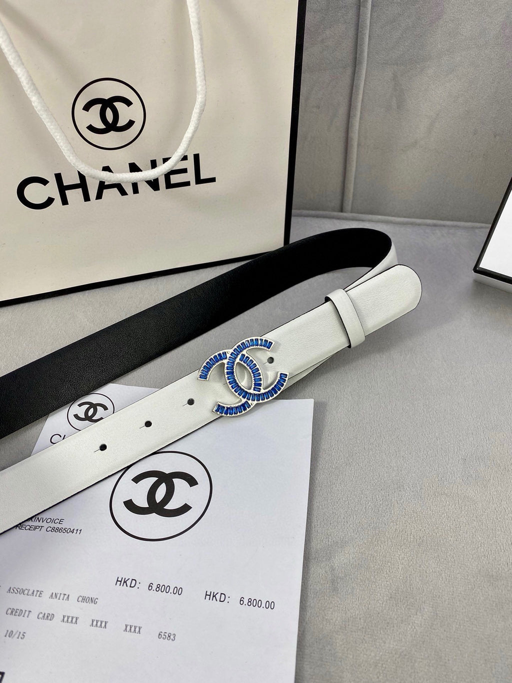 White leather belt with blue CC buckle, resting on a product box.