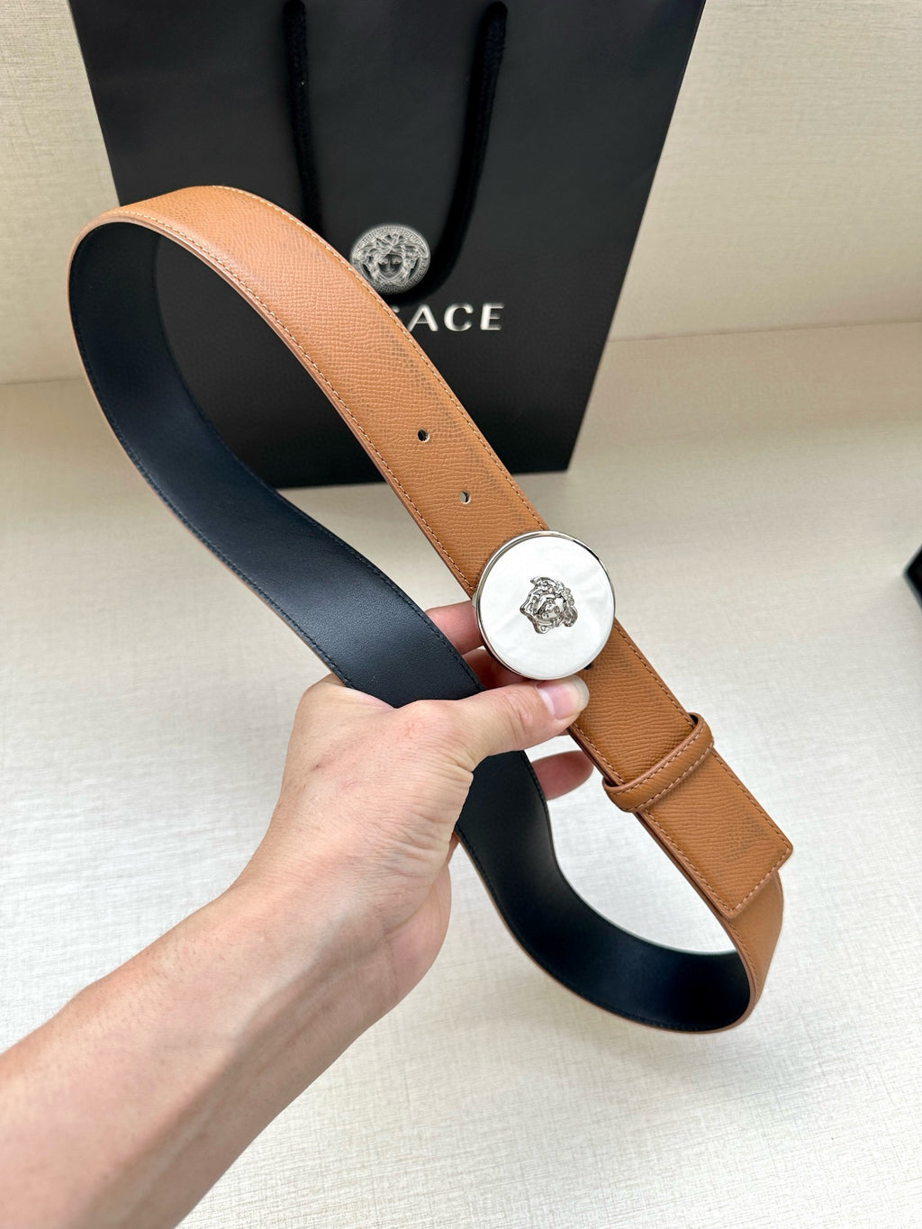 Tan leather belt held up, featuring round silver-toned logo buckle