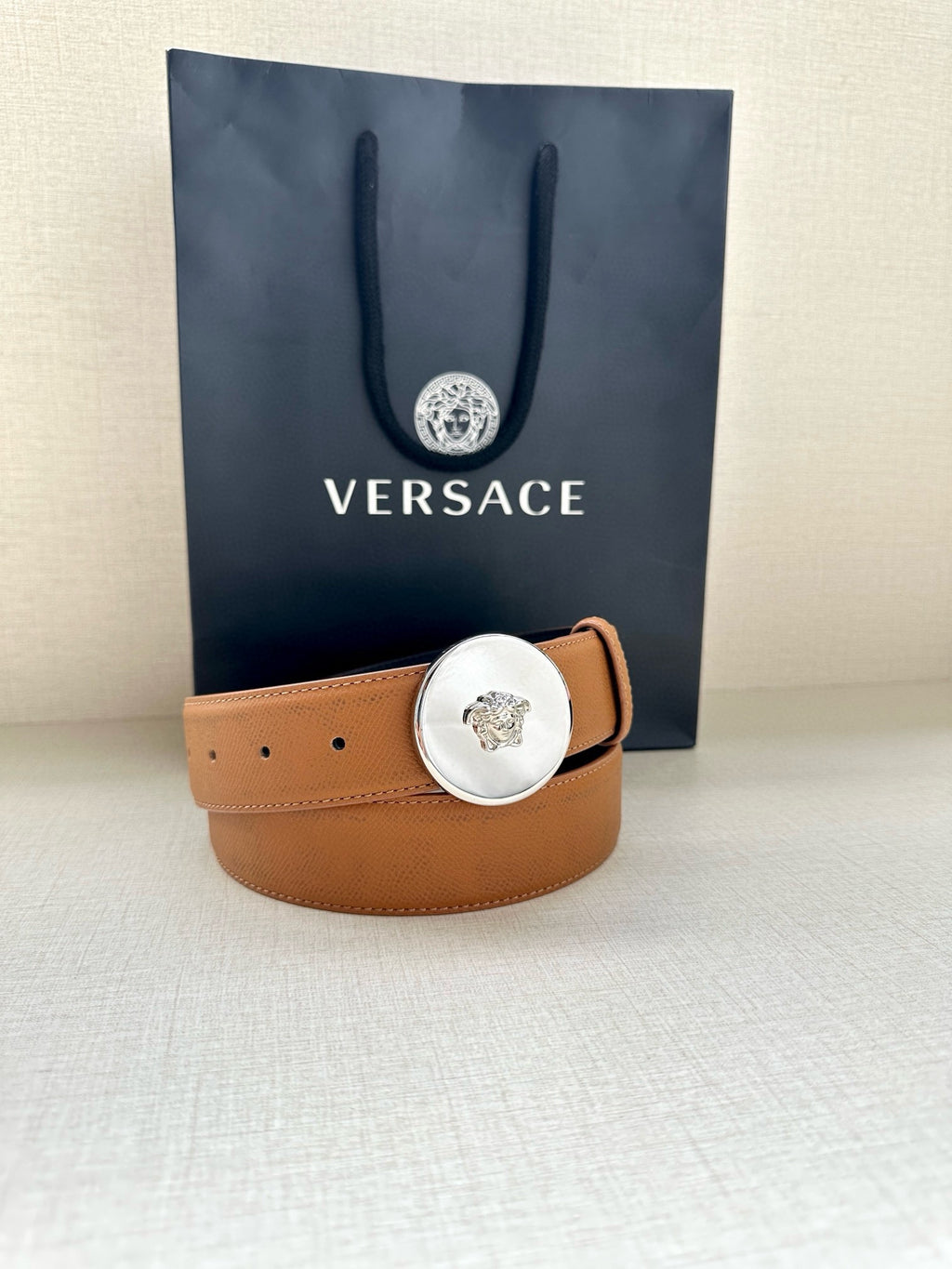 Tan leather belt with round silver-toned logo buckle and short keeper strap
