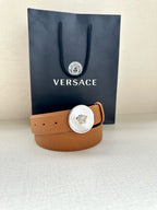 Tan leather belt with round silver-toned logo buckle and short keeper strap