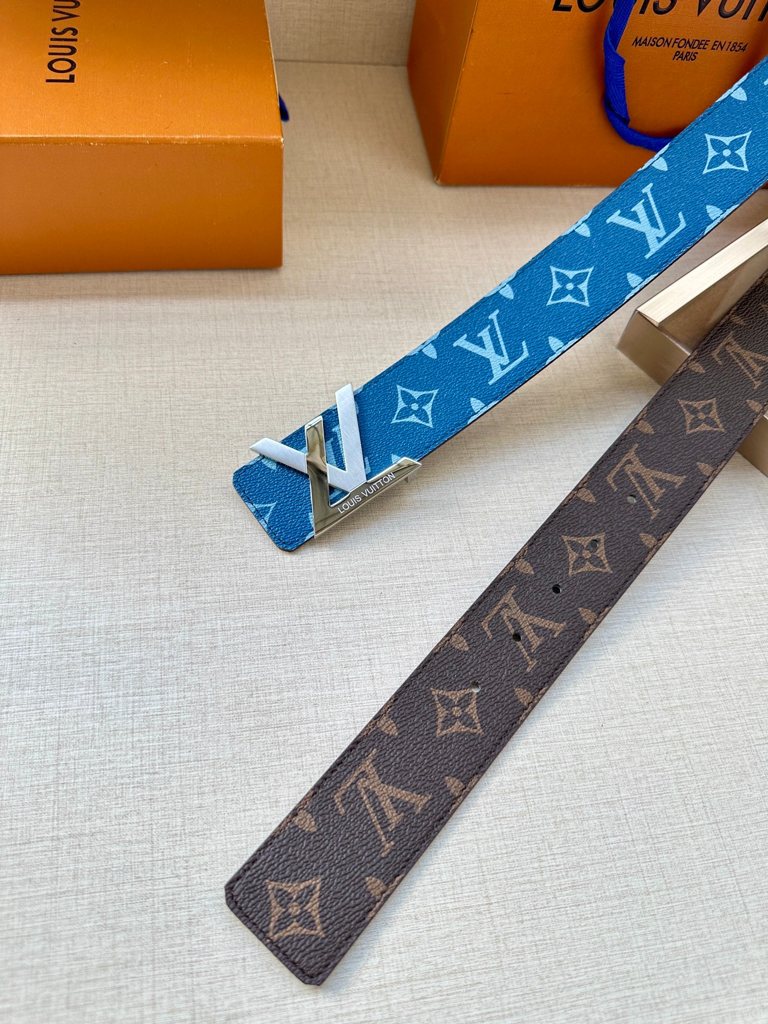 Blue and brown monogram belts with gold-toned buckles, laid on a tray