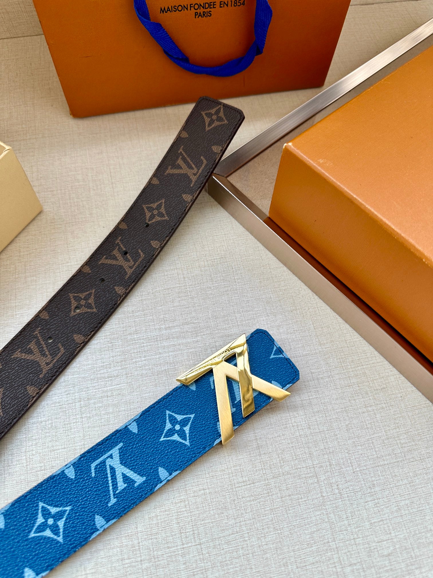 Brown monogram belt strap with gold-toned buckle detail next to blue belt