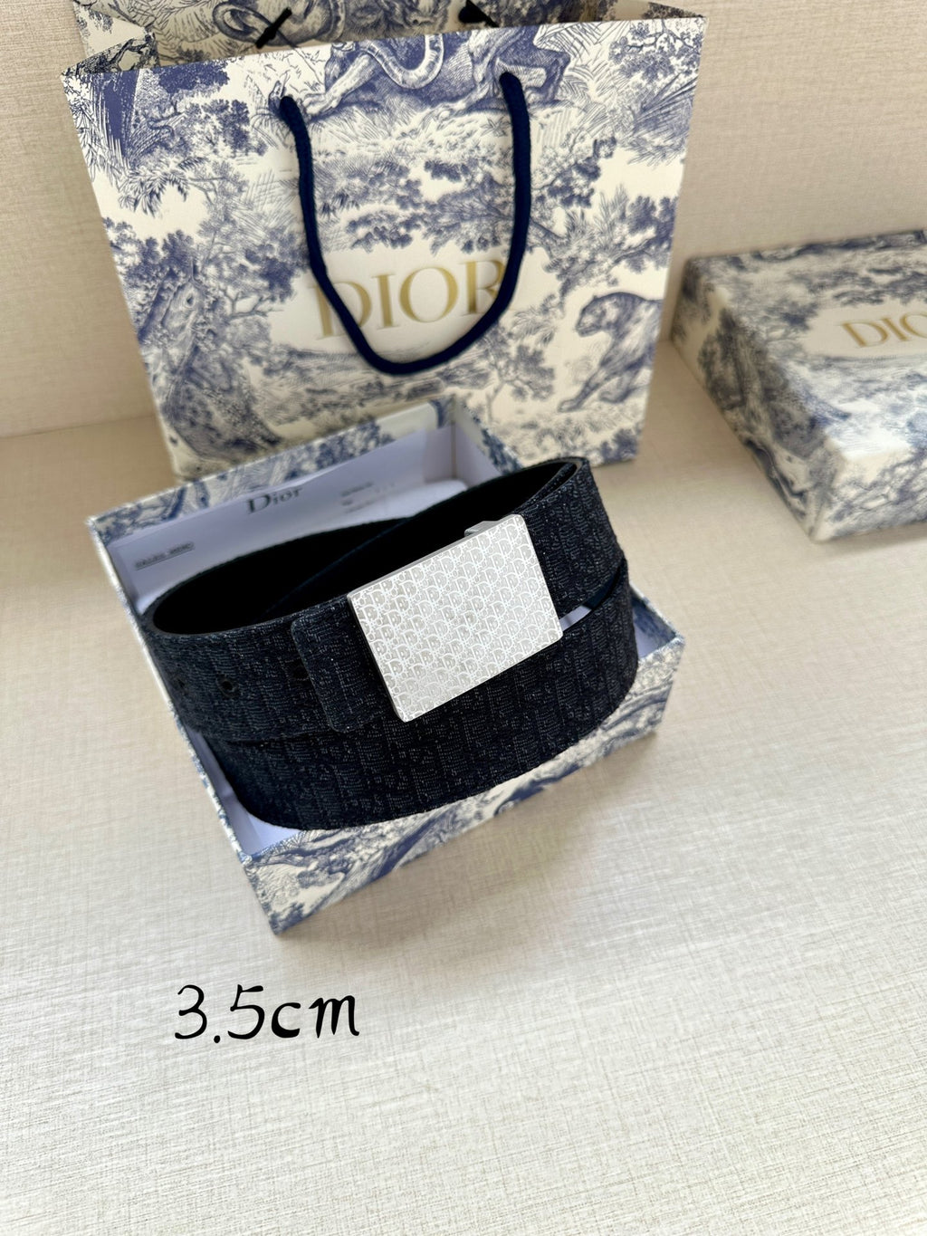 Navy leather belt with silver rectangular logo buckle, displayed with packaging.