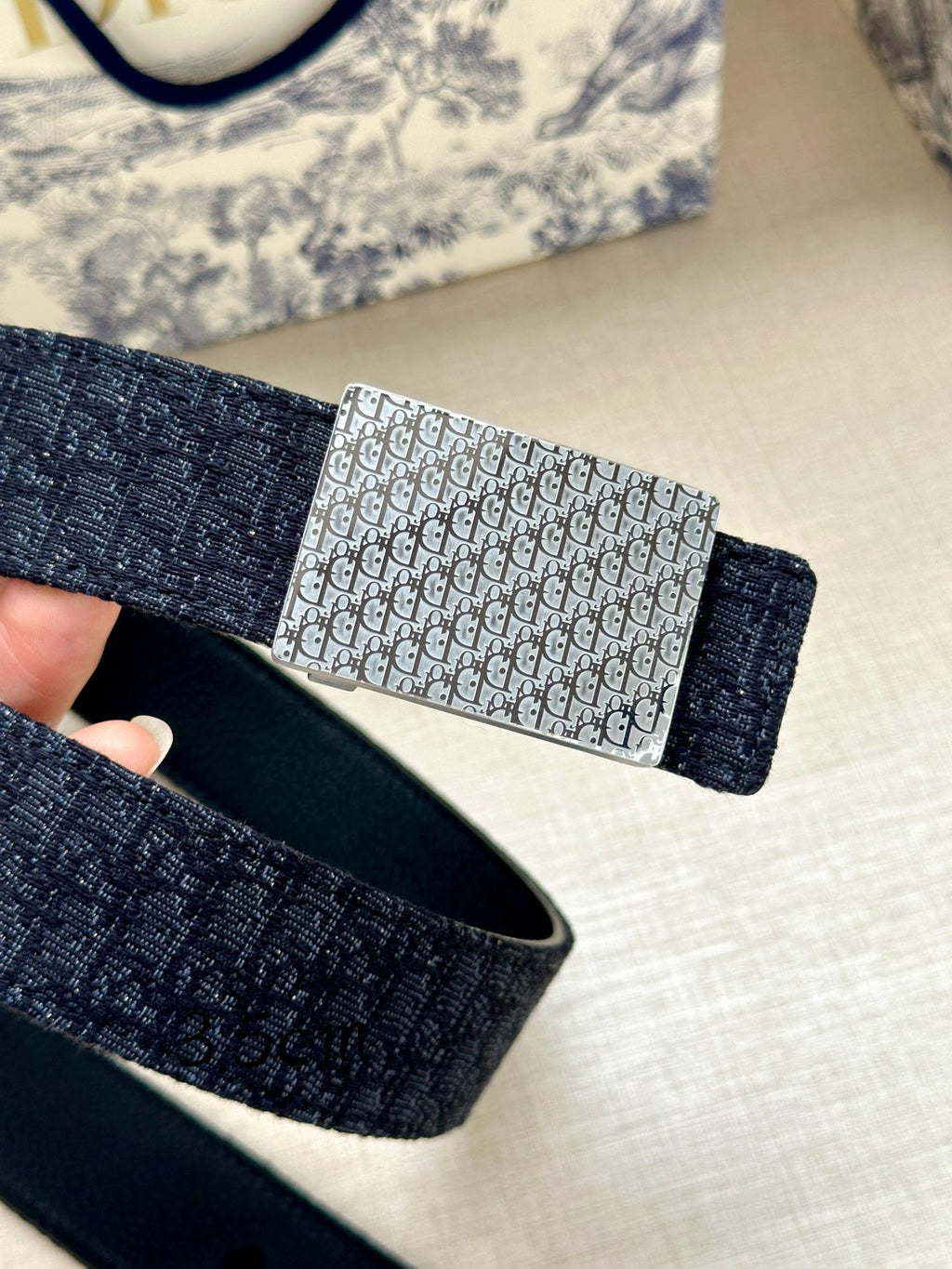 Navy leather belt with silver rectangular monogram buckle, hand holding close-up.
