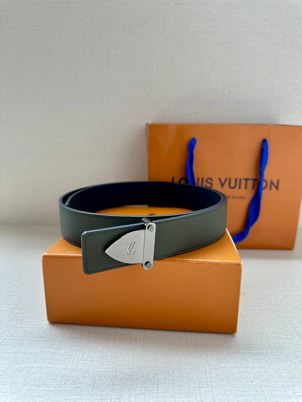 Louis Vuitton leather belt dark green with silver buckle, resting on orange packaging